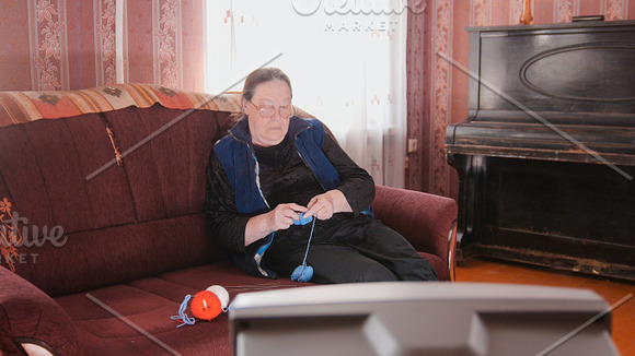 Old lady at home - senior woman watching television and knits wool socks in Graphics