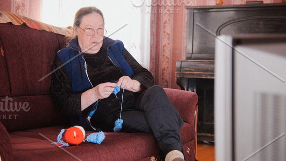 Old lady home at sofa - senior woman watching television and knits wool socks in Graphics