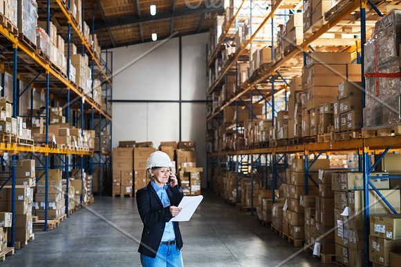 Senior Woman Warehouse Manager Or Supervisor With Tablet And Smartphone Making A Phone Call